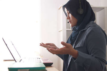 Beautiful Asian muslim woman using headphones speaking her laptop, explaining something. Professional teacher mentor customer service support doing online chat or video call conceptの写真素材