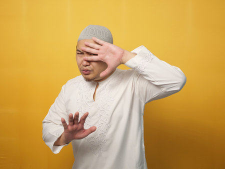 Portrait of funny young  Asian musim man afraid expression with hands raised up, surrender gesture, over yellow backgroundの写真素材