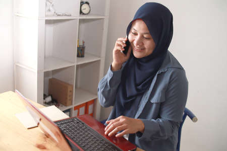 Portrait of happy Asian muslim businesswoman female entrepreneur making business phone call while working at homeの写真素材