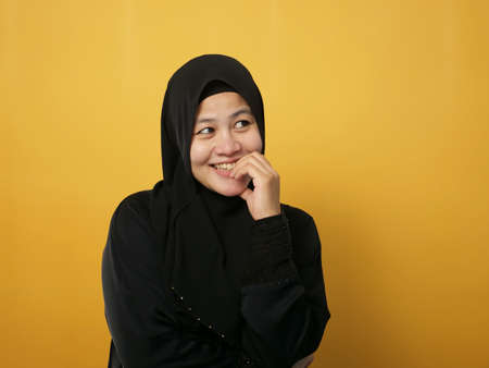 Asian muslim woman wearing hijab shows thinking contemplating expression and smiling happily. Close up portrait against yellow backgroundの写真素材