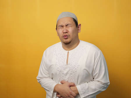 Portrait of young muslim Asian man squezze his stomach for having stomachache pain, against yellow backgroundの写真素材