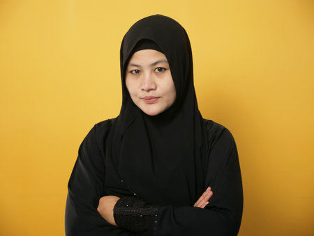 Portrait of Asian muslim woman wearing hijab smiling friendly with crossed arms, confident gesture, successful businesswoman, against yellow backgroundの写真素材