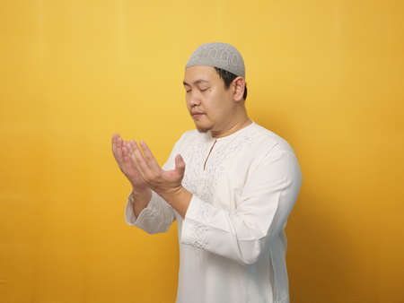 Portrait of Asian muslim man prays to God, praying gesture hands raised up, against yellow backgroundの写真素材