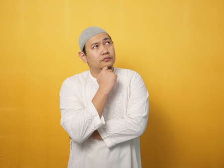 Asian muslim man shows thinking contemplating expression. Looking for solution of a problem. Close up portrait against yellow backgroundの写真素材