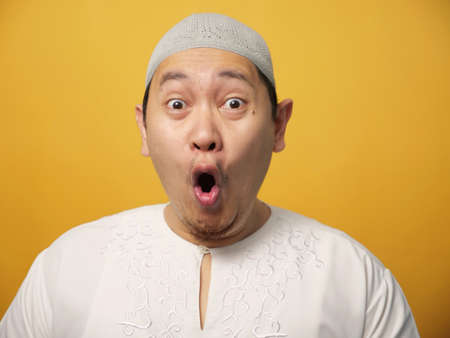 Portrait of cute young Asian muslim man shows surprised or shocked expression with big eyes and open mouth, against yellow backgroundの写真素材