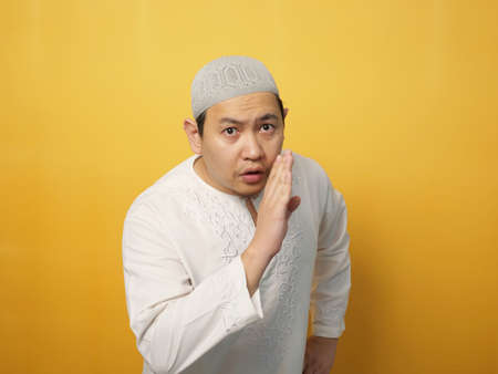 Portrait of young Asian muslim man asking to be quiet, shushing gesture, silence concept, against yellow backgroundの写真素材
