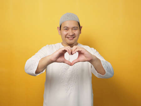 Portrait of Asian muslim man smiling at camera and making love or heart sign gesture with his hands over yellow backgroundの写真素材