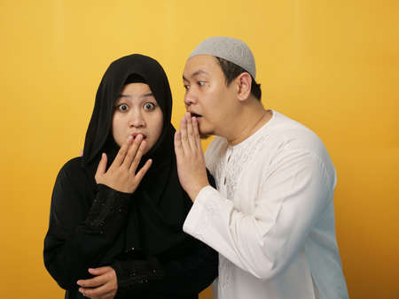 Man covering his mouth and whispering secret to his wife ear, Asian muslim woman looking shocked and excited, opening eyes and mouth widely with surprise, family conceptの写真素材