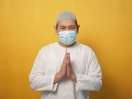 Asian muslim man wearing mask during pandemic new normal, man wearing face protective mask doing greeting gesture, against yellow backgroundの写真素材
