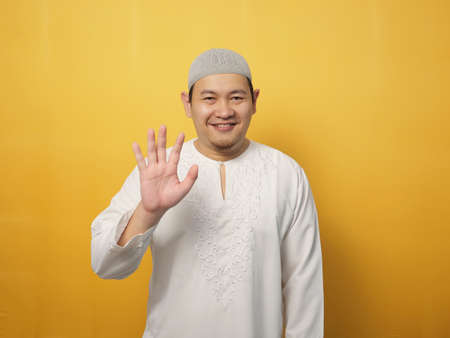 Portrait of muslim man wearing hijab looking at camera smiling and waving his hands saying hi or goodbye farewell gesture against yellow backgroundの写真素材