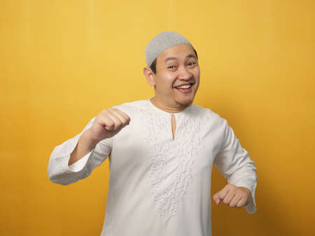 Funny young Asian muslim man dancing happily joyful expressing celebrating good news victory winning success gesture, smiling positive excited emotion, against yellow backgroundの写真素材