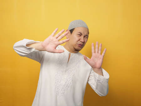 Portrait of funny young  Asian musim man afraid expression with hands raised up, surrender gesture, over yellow backgroundの写真素材