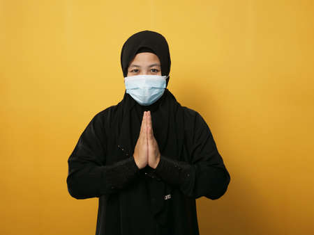 Asian muslim woman wearing hijab and mask during pandemic new normal, woman wearing face protective mask doing greeting gesture, against yellow backgroundの写真素材