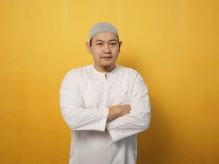Portrait of Asian muslim man smiling friendly with crossed arms, confident gesture, successful businessman, against yellow backgroundの写真素材