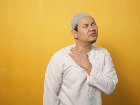 Portrait of young Asian muslim man suffers from neck pain, against yellow backgroundの写真素材