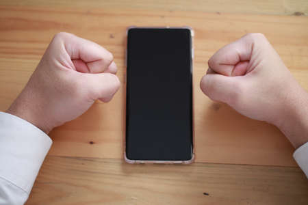 Angry man clasp his fist on table with smart phone, mobile phone mock up conceptの写真素材