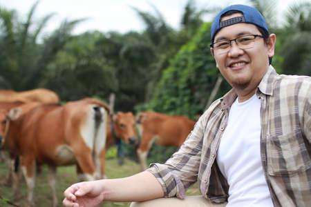 Happy successful Asian farmer looking at camera and smiling, with domesticated cow ox cattle grazing in the background, organic agriculture farming entrepreneurの写真素材