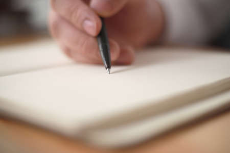 Close up of hand holding pen and writing on empty book paper, selective focus on pen