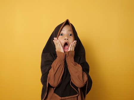 Funny cute Asian muslim little girl wearing hijab looked shocked surprised expression with big eyes and mouth open against yellow backgroundの写真素材