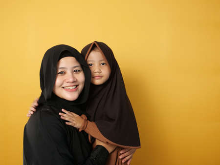 Happy smiling muslim mother and little baby daughter wearng hijab looking at camera against yellow backgroundの写真素材