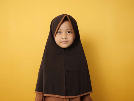 Funny cute Asian muslim little girl wearing hijab looking at camera and smiling, against yellow...
