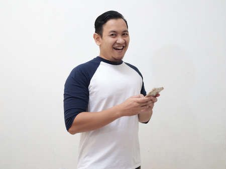 Happy cheerful young Asian man smiling to camera while holding his phone, against white backgroundの写真素材