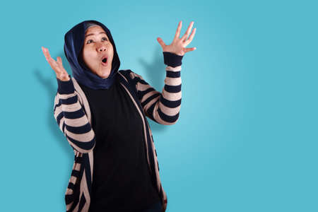 Happy excited Asian muslim woman looking up with hands raised waiting for surprised prize from above, over blue backgroundの写真素材