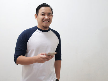 Happy cheerful young Asian man smiling to camera while holding his phone, against white backgroundの写真素材