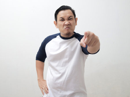 Young Asian man pointing to camera with angry cynical expression, giving warn. Against white backgroundの写真素材