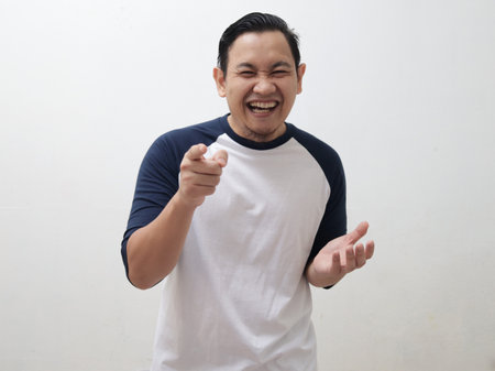 Funny cute Asian man wearing casual shirt smiling and laughing while pointing to camera, against white backgroundの写真素材
