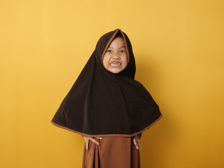 Funny cute Asian muslim little girl wearing hijab looked angry and upset, looking at camera, against yellow backgroundの写真素材