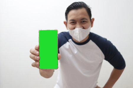 Asian man wearing protective mask shows empty blank green screen on his smartphone, phone mockup templateの写真素材