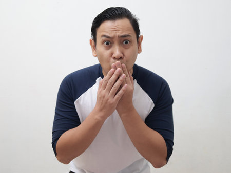 Asian man shocked and worried to see something bad, covering his mouth with hands and big eyes open, over grey backgroundの写真素材
