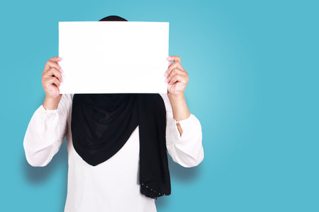 Portrait of muslim woman in hijab covering her face with blank empty paper. copy space mock up for anonymouse person, over blue backgroundの写真素材
