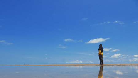Young muslim female wearing hijab standing on the beach and looking at the ocean under bright sun light and blue sky at the background with negative copy spaceの写真素材