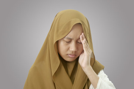 Asian muslim teenage girl wearing hijab holding her head in pain for having headache, close up portrait against grey backgroundの写真素材