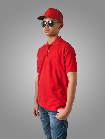 Young Asian teenage boy wearing red collared shirt, hat and sunglasses standing with hands on hip over grey background, t-shirt template mock upの写真素材
