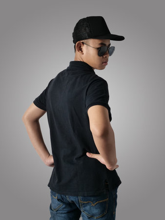 Rear view of Asian boy wearing black shirt hat and sunglasses, looking to the side, against grey...