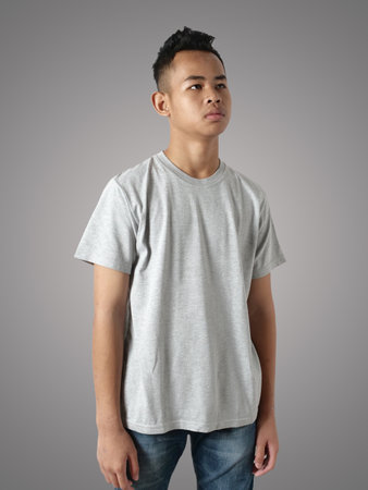 Young Asian teenage boy wearing grey shirt, standing with hands on hip over gray background, t-shirt template mock upの写真素材