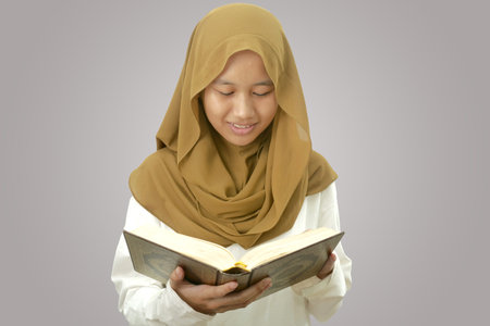 Asian muslim girl wearing hijab holding and reciting Quran, holy book of islam religion, against grey backgroundの写真素材