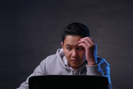Asian man thinking and worried while working on his laptop at midnight, negative emotions in mindの写真素材