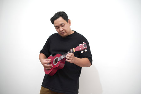Asian man playing ukulele at home, standing against white wallの写真素材