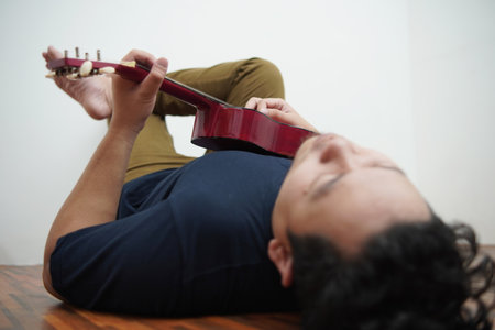 Asian man singing and playing ukulele at home, lying on the floorの写真素材