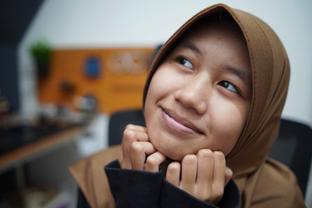 Cute young Asian muslim teenage girl rest her chin on hands, smiling and wondering, happy expression