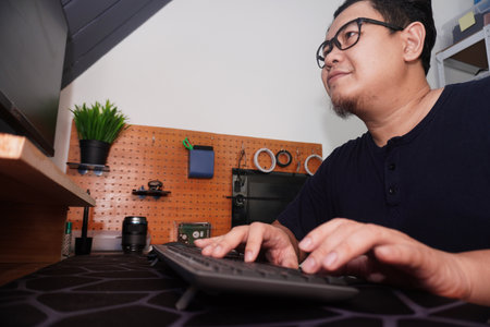 Adult Asian man typing on pc computer at home, freelancer working remotely, online job concept,...