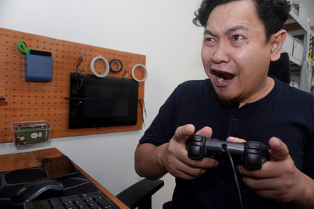 Adult Asian man playing video games on computer, happy smiling expression while holding game console