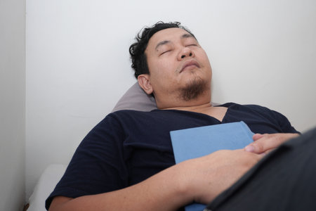 Adult Asian man sleep on bed mattress while holding book, tired relax person reading bookの写真素材