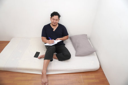 Adult Asian man writing daily journal on his diary on bed before sleep, reflecting his life