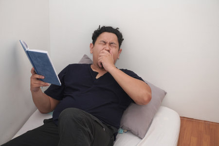 Adult Asian man reading book in bed before sleep, minimalist sparse room with single mattress on floorの写真素材
