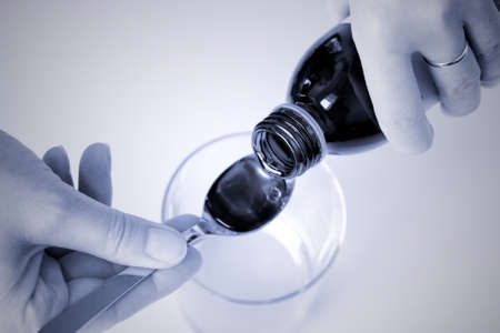 Healthcare concept - close up of a woman's hand pouring cough and sickness syrup into a spoon over a...